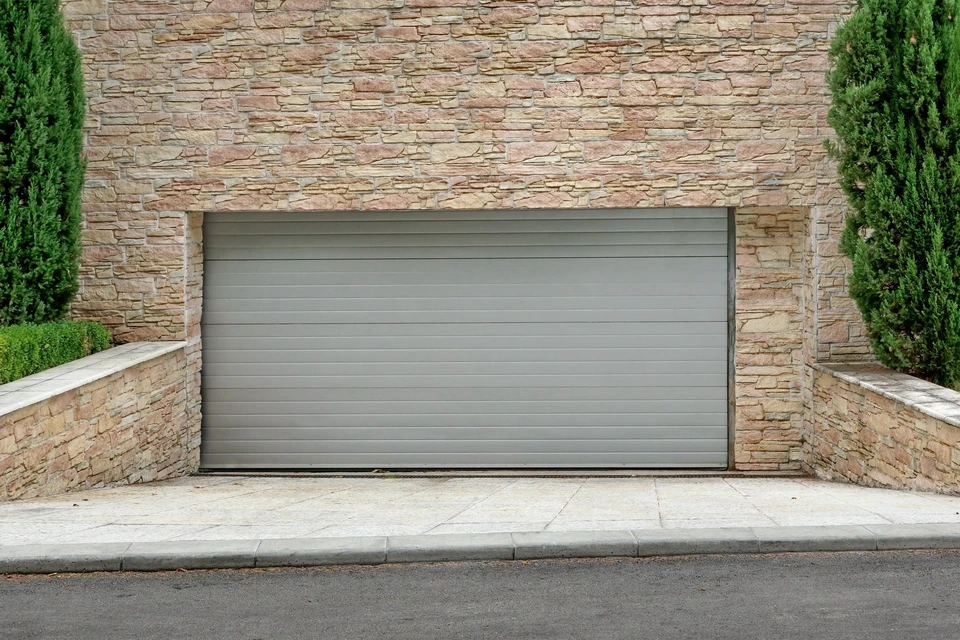 Façade extérieure résidentielle avec porte de garage sectionnelle grise à panneaux horizontaux lisses intégrée dans mur en pierre naturelle beige-rosé