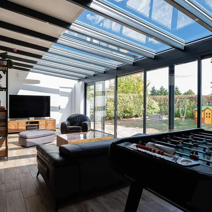 Véranda moderne à structure métallique anthracite avec grandes baies vitrées, adossée à maison traditionnelle aux murs crépis beiges et toiture tuiles canal