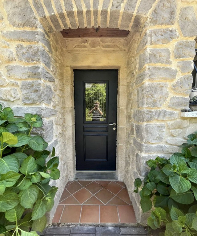 Véranda moderne à structure métallique anthracite avec grandes baies vitrées, adossée à maison traditionnelle aux murs crépis beiges et toiture tuiles canal